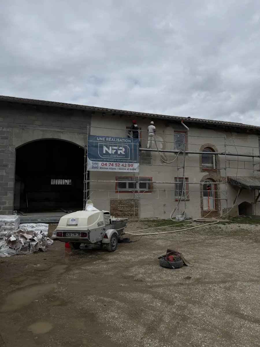 Deux artisans NFR projettent l'enduit sur la façade d'une ferme en pisé, machine à projeter au sol